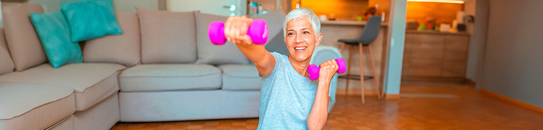 Foto de una señora entrenando con pesas, sentada sobre un tapete de yoga, en la sala de su casa